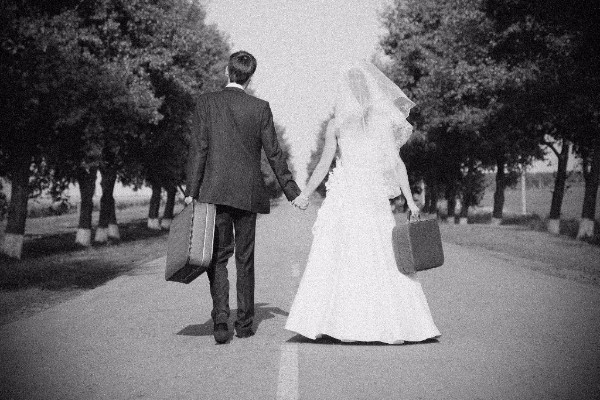 grainy photo of a just married couple walking down road with suitcases in hands